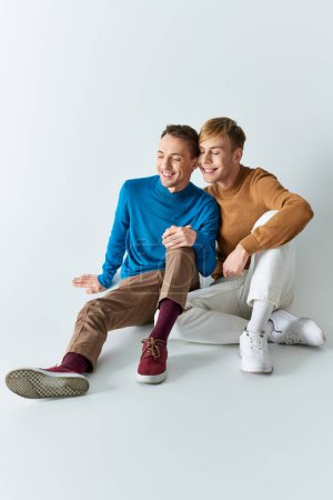 Téléchargez les photos : Un couple gay aimant en tenue décontractée assis étroitement ensemble sur un fond gris. - en image libre de droit