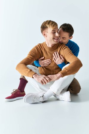 Two men in casual attire sitting on the ground, hugging each other closely.