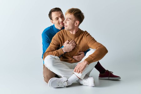 Two men in casual attire sit on the ground with arms around each other.