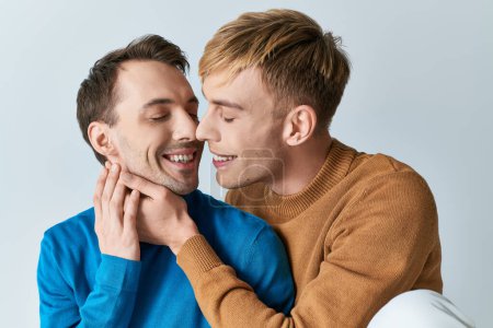 Una pareja gay amorosa con atuendos casuales compartiendo un cálido abrazo en un fondo gris.