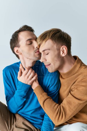 Téléchargez les photos : Un couple gay aimant en tenue décontractée assis l'un à côté de l'autre sur un fond gris. - en image libre de droit