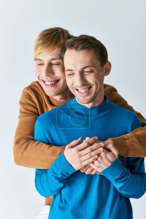 Deux jeunes hommes s'embrassent, souriant joyeusement en tenue décontractée sur fond gris.
