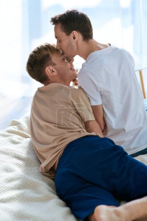 Two men in casual attire sit on a bed, sharing a moment of closeness and peace.