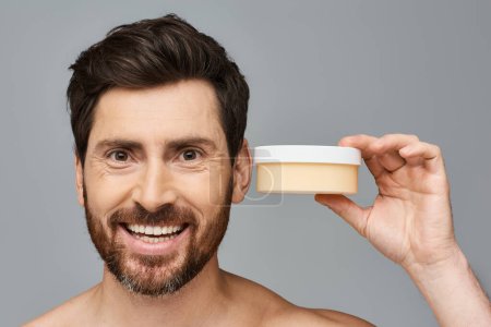 Photo for A man smiling, holding a jar of cream, applying it to his face. - Royalty Free Image