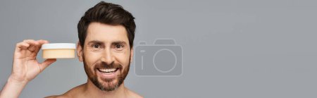 Photo for A bearded man holding a container of cream for skincare. - Royalty Free Image