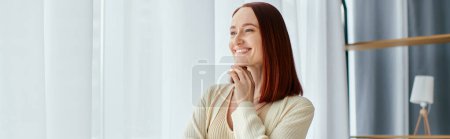 Photo for A red-haired woman smiling by a window, lost in thought. - Royalty Free Image