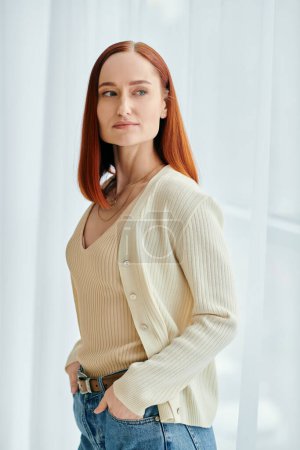 Téléchargez les photos : Une femme rousse en jean et un cardigan frappent une pose devant une fenêtre avec de la lumière naturelle qui brille. - en image libre de droit