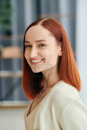 Téléchargez les photos : Une femme aux cheveux cramoisis souriant chaleureusement à la caméra, exsudant bonheur et confort dans un cadre d'appartement moderne. - en image libre de droit
