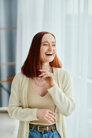 A red-haired woman laughs joyfully in front of a window with the setting sun casting a warm glow.
