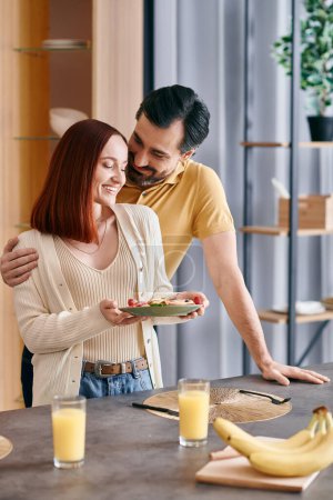 Téléchargez les photos : Une rousse et un homme barbu dégustant un petit déjeuner confortable ensemble dans une cuisine moderne. - en image libre de droit