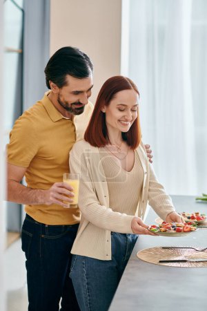 Téléchargez les photos : Une femme rousse et un homme barbu rient et mangent dans une cuisine moderne, créant des souvenirs heureux ensemble. - en image libre de droit