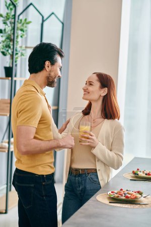 Photo for A beautiful adult couple, a redhead woman and bearded man, standing together in a modern kitchen, enjoying quality time at home. - Royalty Free Image