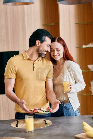Une femme rousse et un homme barbu se tiennent dans une cuisine moderne, tenant une assiette de fruits colorés.