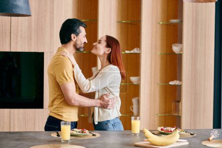 Foto de Una pelirroja y un barbudo comparten un tierno abrazo en una cocina moderna, rodeada de calidez y afecto. - Imagen libre de derechos