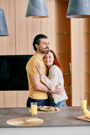 A redhead woman and bearded man embrace in a modern kitchen, sharing a moment of intimacy and affection.