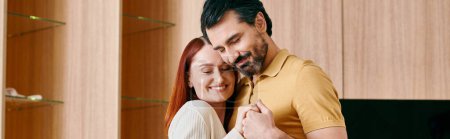 A beautiful adult couple, a redhead woman, and a bearded man, share a heartfelt hug in their modern living room.
