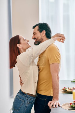 Una pareja adulta, una pelirroja y un barbudo, comparten un tierno abrazo en su cocina moderna, expresando amor y cercanía.