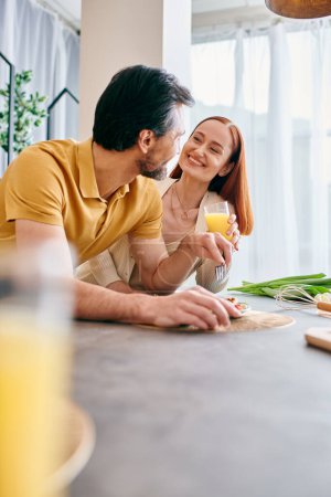 Foto de Una mujer pelirroja y un hombre barbudo se sientan en la mesa de la cocina, saboreando jugo de naranja juntos en su apartamento moderno. - Imagen libre de derechos
