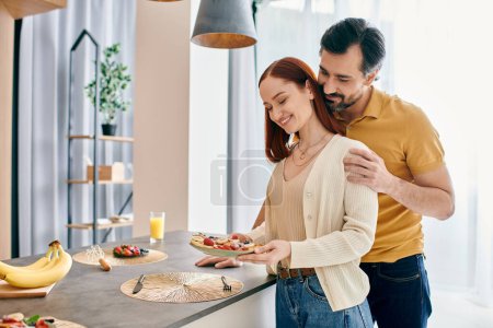 A redhead woman and bearded man stand in a modern kitchen, happily presenting a plate of food they prepared together.