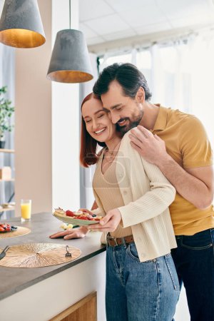 Téléchargez les photos : Une femme rousse et un homme barbu dans une cuisine moderne, tenant une assiette de nourriture délicieuse, profitant de moments de qualité ensemble. - en image libre de droit