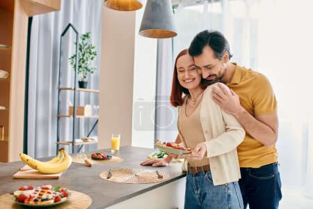 Foto de Un hombre barbudo y una mujer pelirroja disfrutan de un acogedor desayuno juntos en una cocina moderna apartamento. - Imagen libre de derechos