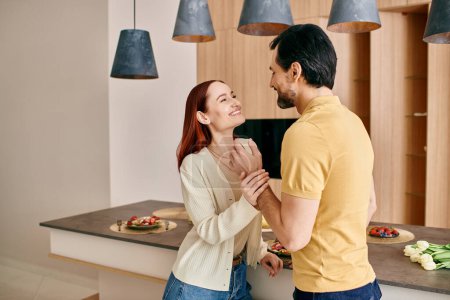A redhead woman and bearded man stand in a modern kitchen, sharing a moment of connection and closeness.