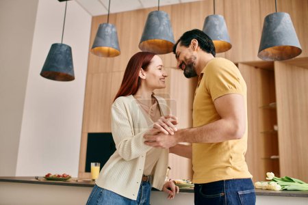 Photo for A beautiful adult couple, a redhead woman and a bearded man, standing together in a modern kitchen, spending quality time. - Royalty Free Image