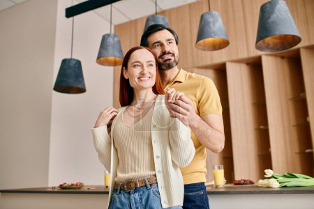 Une femme rousse et un homme barbu se tiennent ensemble dans une cuisine moderne, profitant d'un moment de qualité dans leur appartement.