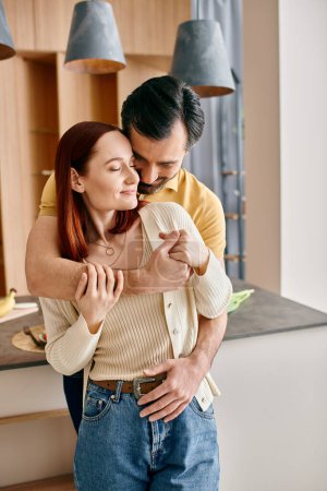 Una pelirroja y un hombre barbudo se abrazan tiernamente en su cocina moderna, compartiendo un momento de amor y conexión.
