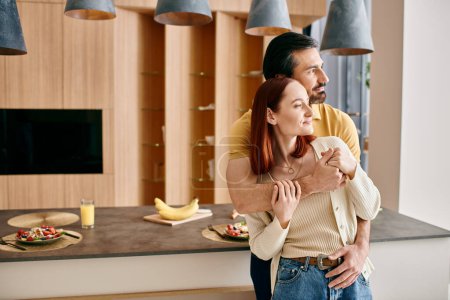 Téléchargez les photos : Femme rousse et homme barbu dans une étreinte chaleureuse dans une cuisine moderne, montrant affection et connexion. - en image libre de droit