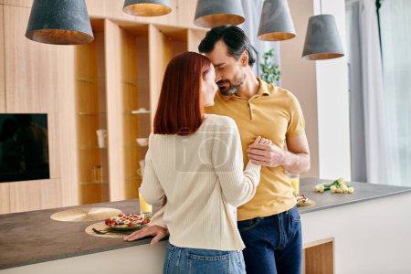 Foto de Una mujer pelirroja y un hombre barbudo abrazan cariñosamente en una cocina moderna, disfrutan de tiempo de calidad juntos - Imagen libre de derechos