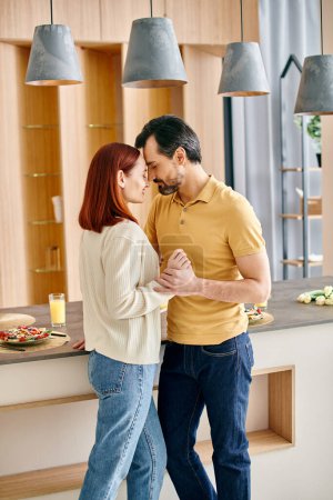 A bearded man and a redhead woman are happily dancing together in a sunlit modern kitchen.