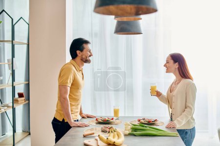 Photo for A redheaded woman and bearded man prepare a meal together at the kitchen counter of their modern apartment. - Royalty Free Image