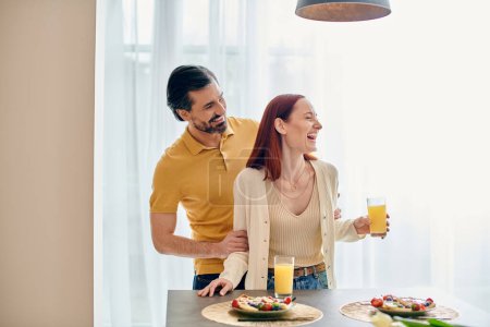 Foto de Una pelirroja y un hombre barbudo disfrutan del desayuno juntos en un apartamento moderno, conectando comida y conversación. - Imagen libre de derechos