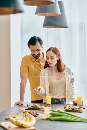 Foto de Una pelirroja y un barbudo se sientan a la mesa de la cocina, riendo y disfrutando del desayuno juntos en un apartamento moderno. - Imagen libre de derechos