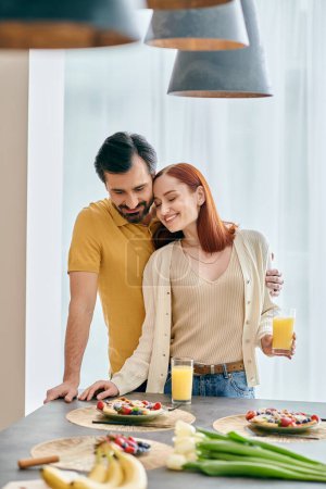 Photo for A beautiful adult couple, a redhead woman and a bearded man, strike a pose in a modern kitchen with food on the counter. - Royalty Free Image