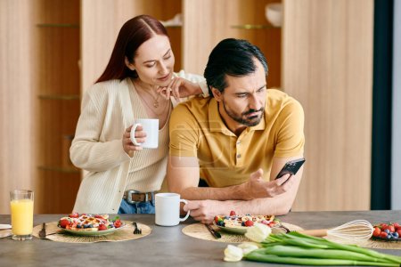 Foto de Una pelirroja y un hombre barbudo absortos en sus teléfonos mientras comparten el desayuno en un apartamento moderno. - Imagen libre de derechos