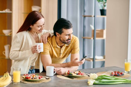 Una mujer pelirroja y un hombre barbudo disfrutan del desayuno mientras están absortos en sus teléfonos en un entorno de apartamento moderno.