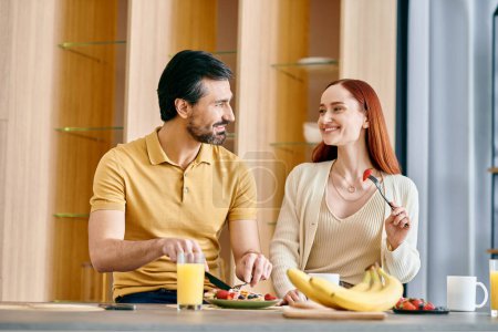 Foto de Un hombre barbudo y una pelirroja disfrutan de un acogedor desayuno juntos en una cocina moderna, creando una escena cálida y armoniosa. - Imagen libre de derechos
