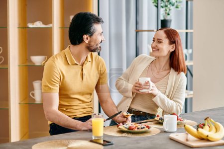 A bearded man and a redhead woman enjoying breakfast together at their kitchen table in a modern apartment.