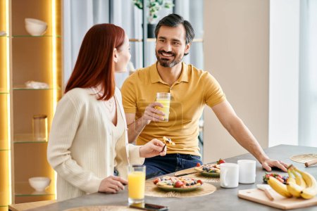 Téléchargez les photos : Une femme aux cheveux roux et un homme barbu aiment passer du bon temps ensemble, savourant du jus d'orange à leur table de cuisine. - en image libre de droit