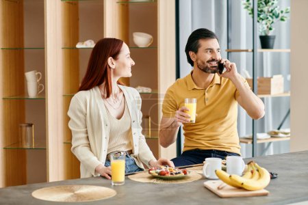 Foto de Redhead woman and bearded man chatting on phone while enjoying breakfast in modern apartment. - Imagen libre de derechos