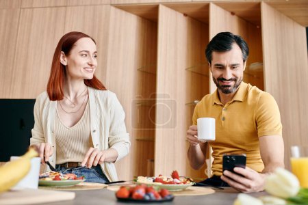 Foto de Una mujer pelirroja y un hombre barbudo se sientan en una mesa, desayunando felizmente en su apartamento moderno. - Imagen libre de derechos