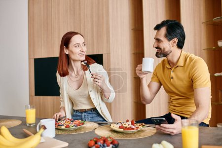 Une rousse et un barbu sont assis à une table de cuisine, mangeant joyeusement le petit déjeuner ensemble dans un appartement moderne.