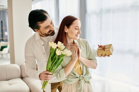 Téléchargez les photos : Un homme barbu présente tendrement un bouquet de tulipes à une rousse dans un cadre d'appartement moderne, incarnant amour et convivialité. - en image libre de droit