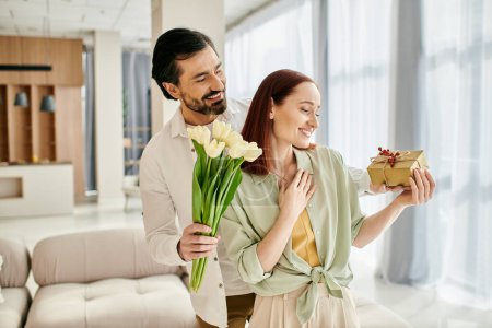 Photo for A beautiful adult couple, a redhead woman, and a bearded man, standing in a modern living room holding a gift box. - Royalty Free Image