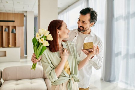 Téléchargez les photos : Une femme rousse et un homme barbu tenant une boîte-cadeau, partageant un moment spécial dans leurs appartements modernes salon confortable. - en image libre de droit