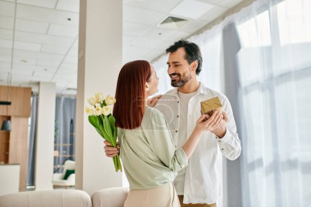 Photo for A man with a beard is giving a gift to a beautiful redhead woman in a stylish living room. - Royalty Free Image