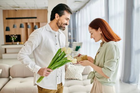 Foto de Un hombre barbudo está entregando flores a una pelirroja en su moderna sala de estar. La pareja está disfrutando de un momento de corazón juntos. - Imagen libre de derechos