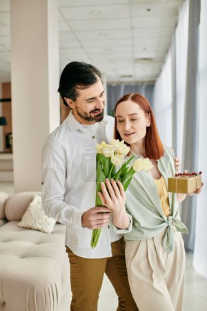 Foto de Una mujer pelirroja y un hombre barbudo sostienen flores en una sala de estar moderna, disfrutando de un tiempo de calidad juntos. - Imagen libre de derechos
